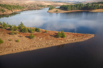 : High-angle view of a calm reservoir reflecting the sky, bordered by forested hills and a rocky shoreline. Captures a peaceful, expansive natural landscape scene.