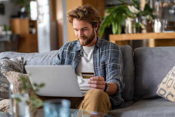 A young White man sitting on the sofa at home in a living room with a laptop on the lap and holding a credit card. Online shopping, payment, purchase, buy. E-commerce.