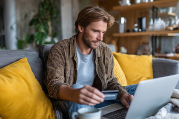 A young White man sitting on the sofa at home in a living room with a laptop on the lap and holding a credit card. Online shopping, payment, purchase, buy. E-commerce.
