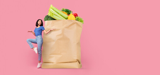 A customer stands next to a large paper bag brimming with fresh vegetables like lettuce, broccoli,...