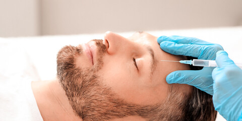 Beautician making face lifting procedures for handsome middle aged man at aesthetic clinic, side view. Man getting beauty injection against forehead wrinkles from unrecognizable plastic surgeon