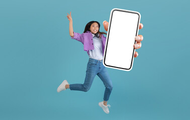 Excited asian lady showing empty smartphone screen while jumping up over blue studio background, collage, full body length, banner. Happy woman recommending cool mobile app or website © Prostock-studio