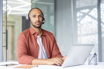 Obraz premium A focused call center agent, wearing a headset, looks at the camera while sitting at a desk. The man works on a laptop in an office environment.
