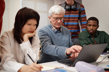 Closeup of professor pointing at laptop screen with students watching and taking notes in college classroom