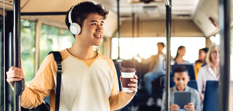 Youth Lifestyle. Positive asian guy standing in public transportation, wearing headphones holding takeout cup and handle, drinking coffee, riding to university looking out of window, banner, panorama