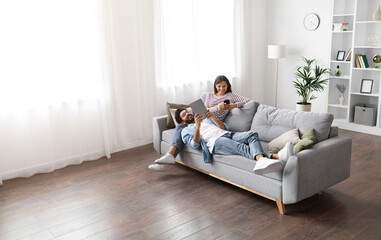 Gadget addiction, modern technologies and millennials lifestyle. Beautiful mixed race spouses resting on sofa at home, using digital tablet and smartphone, high angle view, copy space