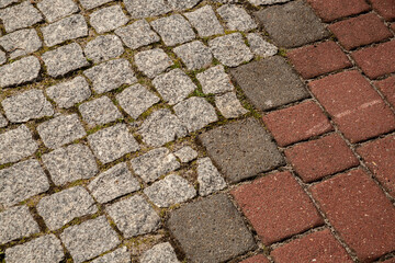 Close-up of a textured transition between grey granite cobblestones and red concrete paving blocks, showing detailed urban surface pattern with moss in between