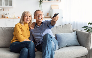 Happy Senior Spouses Watching TV Pointing Television Remote Control Sitting And Hugging On Sofa At...