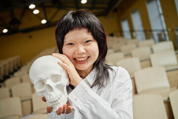 Vertical portrait of young Asian woman as med student posing with human skull model in university...