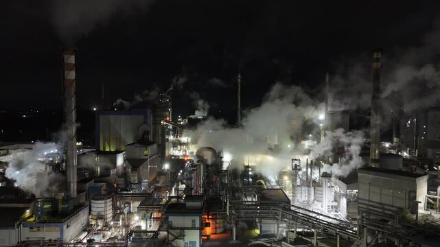 Complexo industrial iluminado &agrave; noite com fuma&ccedil;a das chamin&eacute;s. A&ccedil;&atilde;o cont&iacute;nua e estrutura moderna de uma ind&uacute;stria de grande porte