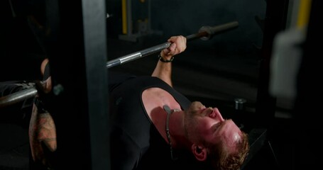 man is focused on lifting a heavy barbell during bench press exercises. He is wearing headphones and is training in a well equipped gym environment.