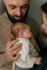 A couple lovingly holds their newborn baby in a cozy indoor setting. The mother gazes at the baby while the father supports them, showcasing a moment of tenderness and family bonding.