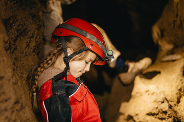 A 6-year-old girl in a protective suit and helmet with a flashlight in the middle of a natural underground cave is walking the tourist route of a caver
