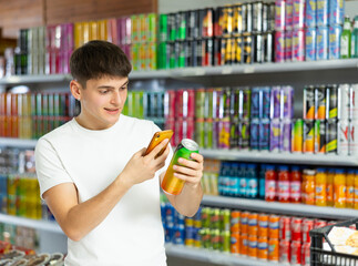 Young guy stands in drinks section of supermarket and uses his phone to find out information about...