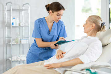 Fototapeta premium Positive young woman doctor conducting questionnaire of elderly female patient lying on couch in medical office, asking series of questions and noting down responses in document folder