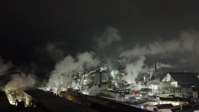 produ&ccedil;&atilde;o de papel e celulose em opera&ccedil;&atilde;o. F&aacute;brica iluminada, chamin&eacute;s ativas e estruturas industriais em destaque sob o c&eacute;u escuro, vista a&eacute;rea de drone