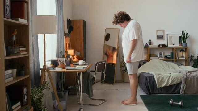 Wide shot of stocky curly haired man in comfy outfit stepping on digital scale in bedroom with wooden furniture and green plants