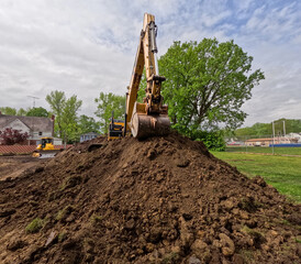 An industrial excavator, bulldozer or backhoe uses a hydraulic arm with a scoop to move dirt and grass in a field. City location with cloudy blue skies.