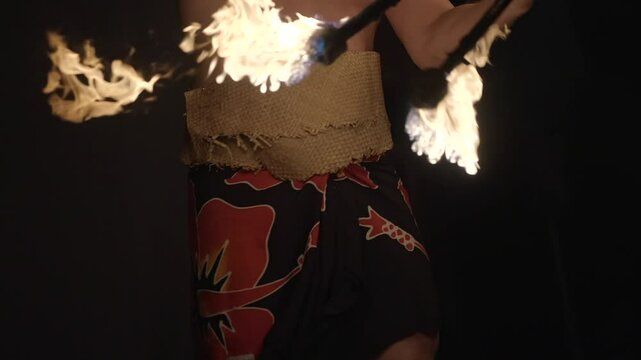 A dynamic midsection view of a male fire dancer skillfully twirling a flaming torch against a dark night sky. The swirling fire creates vivid light trails and dramatic contrast.