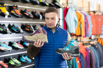 In store, man hold examine several copies item of winter comfortable hiking shoes in hands and...