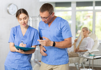 Obraz premium Male doctor and female nurse are standing in medical office with documents and talking about current situation, discussing patiens recovery process. Blurry client in background