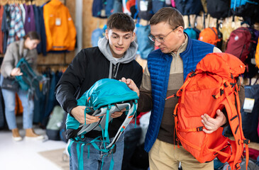 Fototapeta premium Guy with father chooses autumn backpack in store for long hikes and climbing mountains. Travel accessories of different price categories.