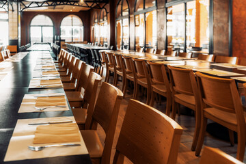 Spacious empty dining hall with long wooden tables, neatly arranged wooden chairs, place settings with napkins, forks, and knives, and large arched windows in warm lighting; wide-angle shot