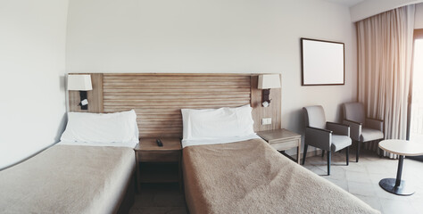 Wide-angle fisheye view of a modern hotel room with two single beds, neutral beige bedding, wooden headboard, bedside tables, two armchairs, curtain-covered window, and natural sunlight
