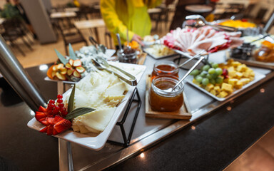 Elegant breakfast buffet with assorted cheeses, fresh sliced fruits, cold cuts, jams in glass jars, and decorative garnishes, arranged on white plates with serving utensils