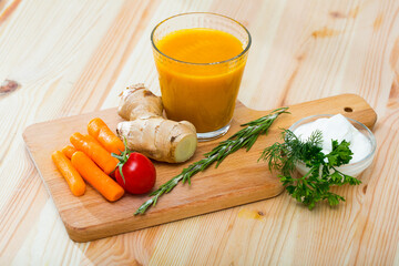 Close up of glass with carrot juice with ginger, greens and vegetables on wooden desk
