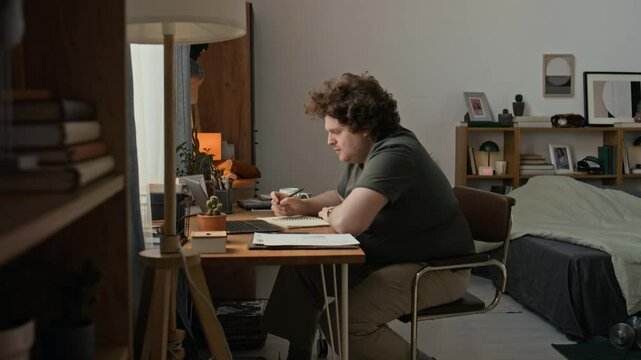 Stocky man sitting at wooden table with notepad and laptop on tp while working remotely at home