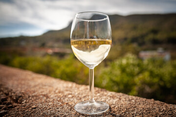 Dewy glass of chilled Spanish white wine stands on a stone terrace; blurred Mediterranean hills and villages create a relaxed holiday ambience under warm afternoon sun.