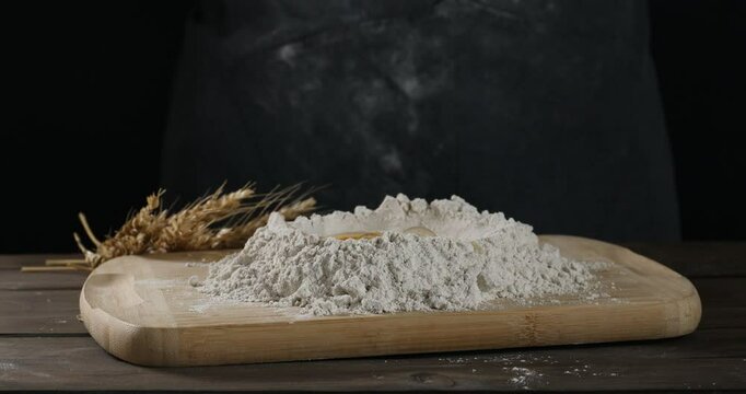 Adding egg into pile of flour at wooden table against black background, closeup. Making dough
