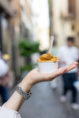 Delicious scoop of gelato held by a hand in a vibrant street with people and greenery around