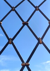 black iron fence with diamond pattern stretches across view, set against vivid blue sky. sunlight reflects off metal, emphasizing its sturdy design and craftsmanship. close up.