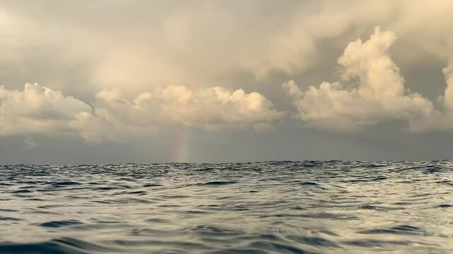 Textured ocean waves rolling beneath a beautifully moody cloudy sky with a distant rainbow. Captured in slow motion from within the ocean at sea level.