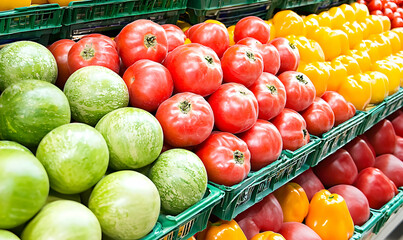 Colorful produce display green melons, red tomatoes, and yellow bell peppers arranged neatly on shelves, in a grocery or market setting