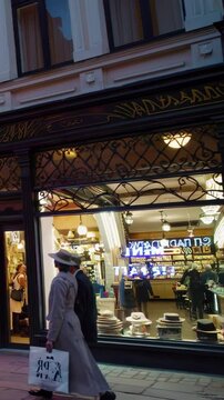 Vertical screen, stylish couple walking past illuminated hat shop, carrying shopping bags while strolling through vibrant city evening streetscape