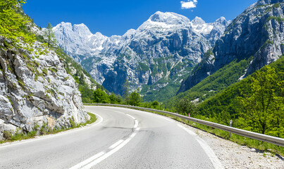 Naklejka premium Scenic mountain road curves through lush green hills towards snow-capped peaks under a bright blue sky