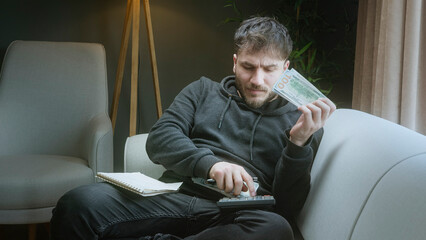Stressed man calculating his remaining money and expenses at home, holding dolar banknotes and...