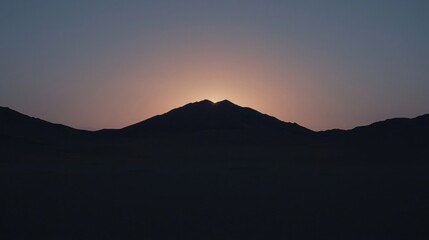 Fototapeta premium A silhouette of a mountain at sunset with the sun glowing just behind the peak against a darkening sky.