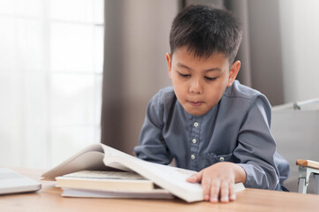 Asian young boy reading book at wooden table in bright room, showing concentration and curiosity. soft lighting enhances peaceful atmosphere