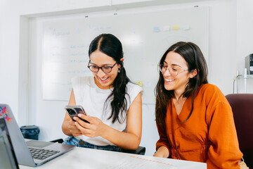 Young Latina women, and coworkers, look at the phone together and smile