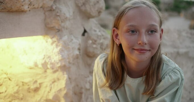 Young blonde girl in light green shirt standing near illuminated stone wall, leaning forward while gazing directly at camera during golden hour lighting, capturing casual adolescent portrait