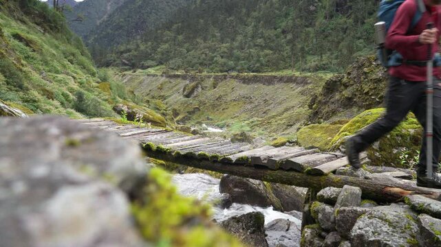 Man with backpack and trekking poles crossing mountain creek wooden bridge during Makalu Barun National Park trek in Nepal. Mountain hiking and active people concept image.