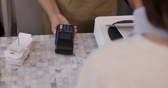 Female barista in brown apron holding credit card terminal while customer hand inserts blue card during payment process in cafe, motion indicates quick transaction and efficiency