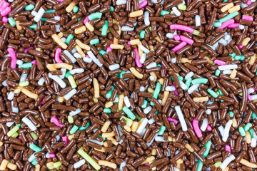 Top view shot reveals a dense scattering of colorful sprinkles. The majority of the sprinkles are chocolate brown, interspersed with vibrant hues of pink, white, light green, and orange.