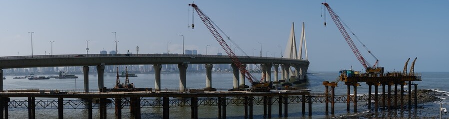 Fototapeta premium Wide Panoramic View of the Bandra-Worli Sea Link Stretching Over the Arabian Sea in Mumbai, India, Framed by Cranes and Pier Structures in the Foreground