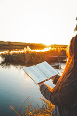 Woman reading open Bible by a peaceful lake during golden sunset, with warm light reflecting on...