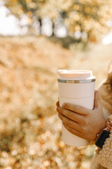 The girl holds a white thermos cup in her hands and enjoys the beautiful autumn